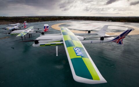 This is an Image of a group of small airplanes/drones together on a helipad the closest to the camera has green and yellow wings with NHS logo Photo credit: Image Credit: Apian